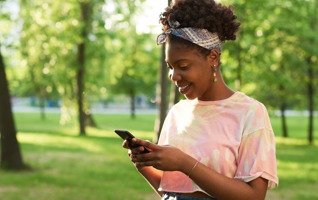 African young woman reading a message on mobile phone and smiling while standing outdoors