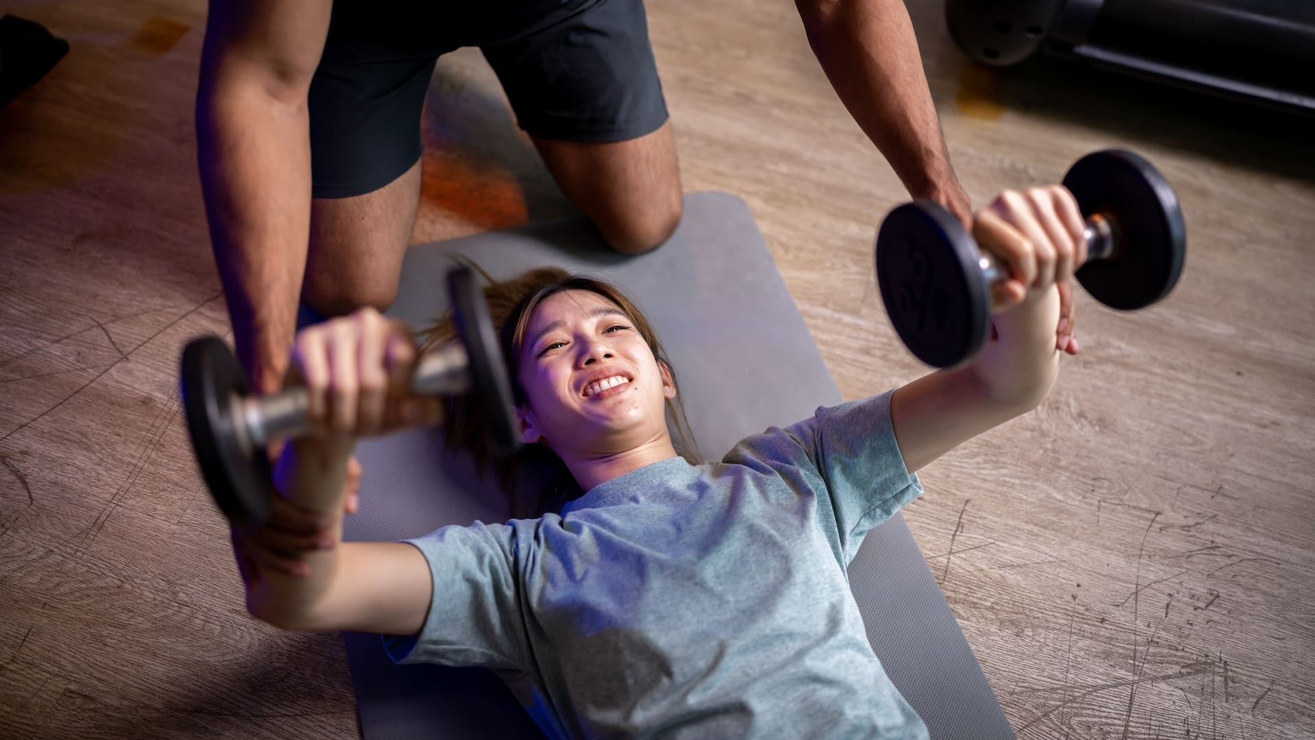 Female student lying on a gym mat doing bicep raises with the help of a spotter