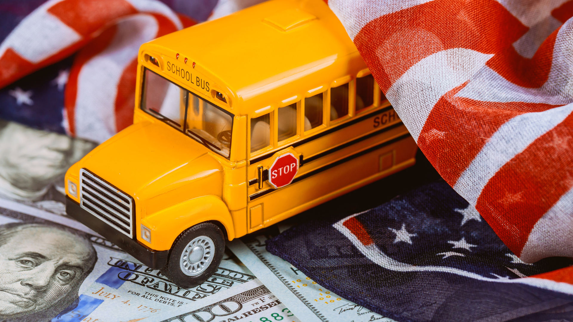 Bus flag cash Close-up of a toy school bus under an American flag with US banknotes scattered beneath it.