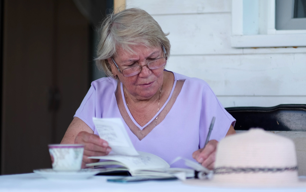 An elderly woman checks receipts against a notebook with a pen in her hand.