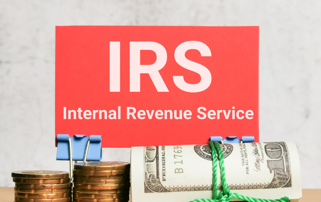 IRS Internal Revenue Service written in white on a red card behind two stacks of pennies and a roll of cash.
